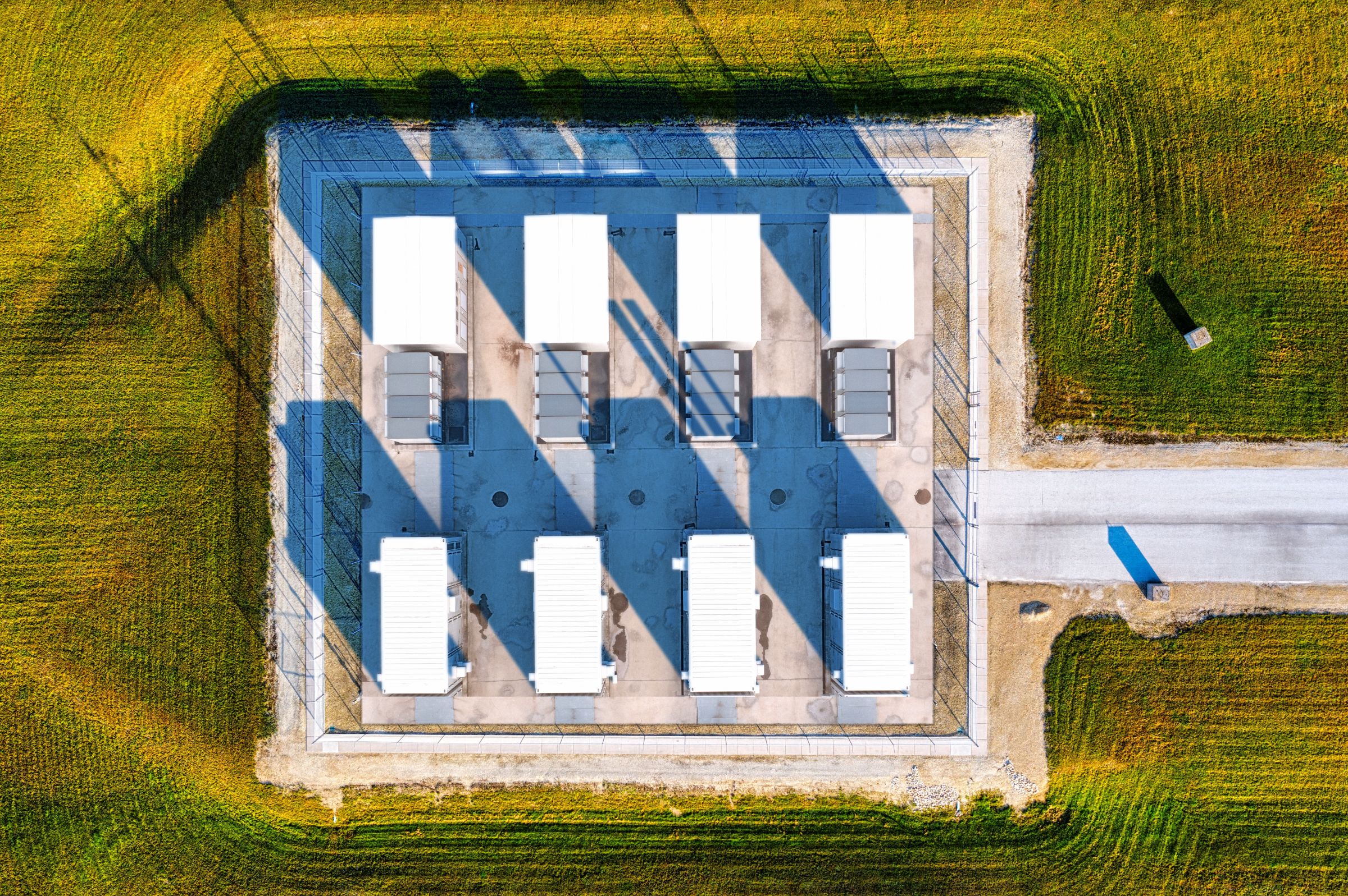 Vue aérienne d’un petit site de stockage par batteries : des conteneurs blancs sur une dalle clôturée, avec son chemin d’accès, au milieu d’une grande parcelle enherbée